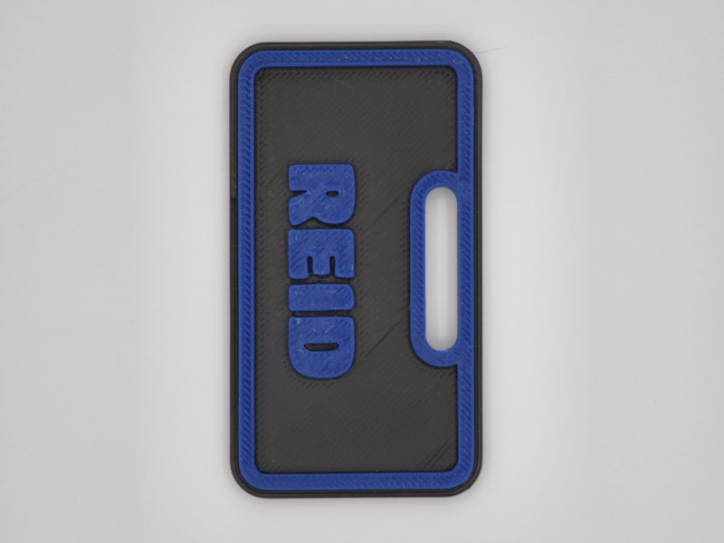 3D-printed REID backpack tag, blue raised bubble letters and border on a black plate with a strap slot